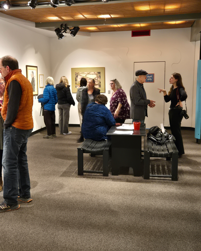 People in an art gallery viewing artwork. Some stand, others interact. A desk sits in the foreground.