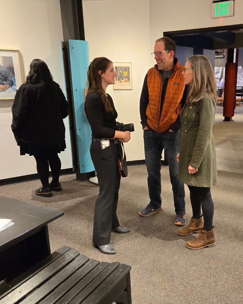People converse in an art gallery, a woman takes a photo.