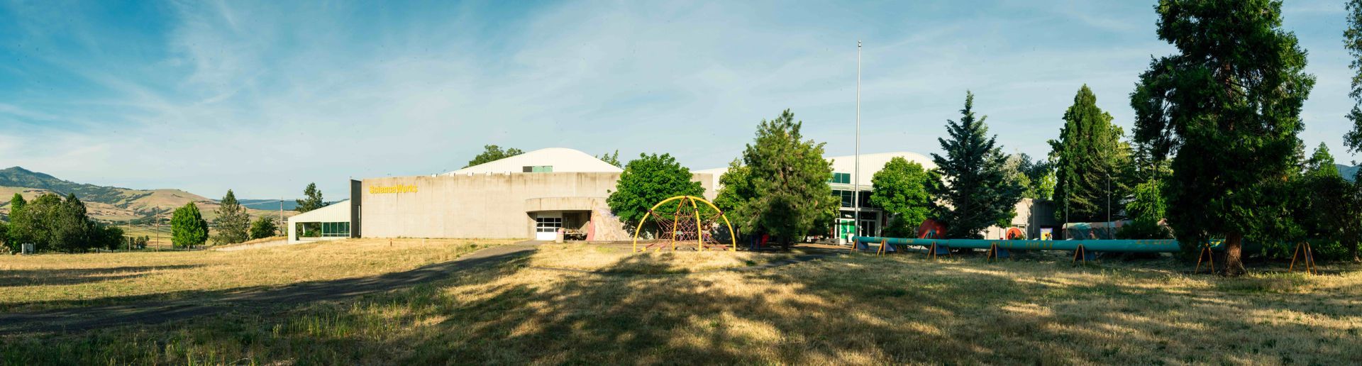 A long, one-story concrete building sits in a grassy field under a bright blue sky, surrounded by green trees.
