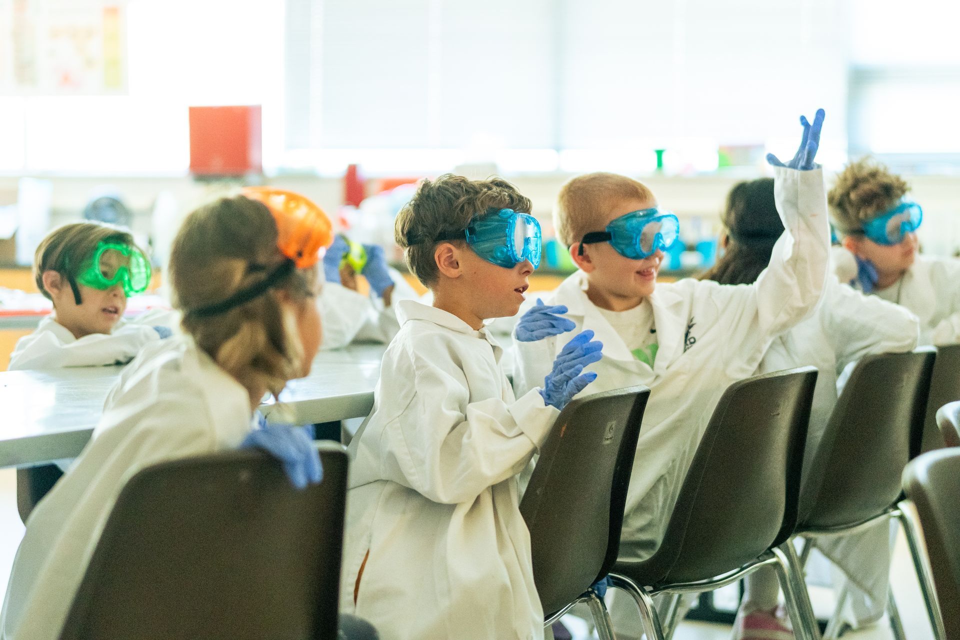 Campers in lab coats, gloves, and goggles watch an educator in a classroom at ScienceWorks Museum