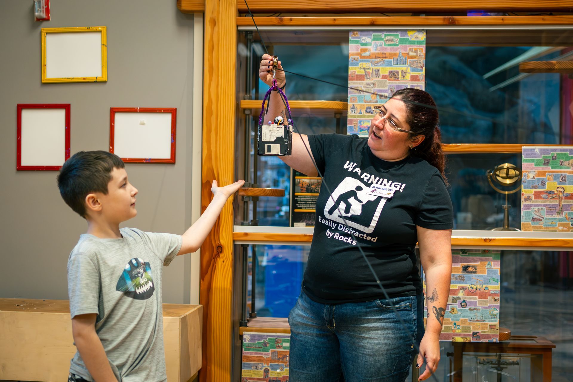 Woman showing object to boy; both in a room with posters, wooden features, and a window.