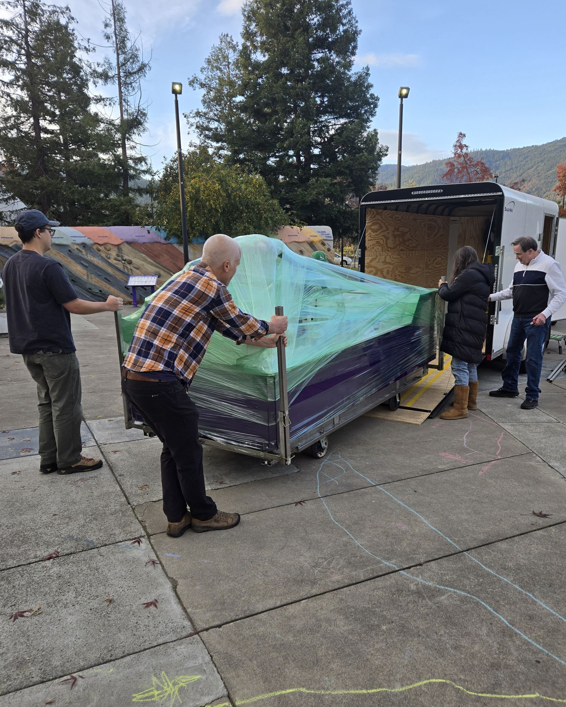 Four people unloading the new water table exhibit, wrapped in plastic, from the trailer.