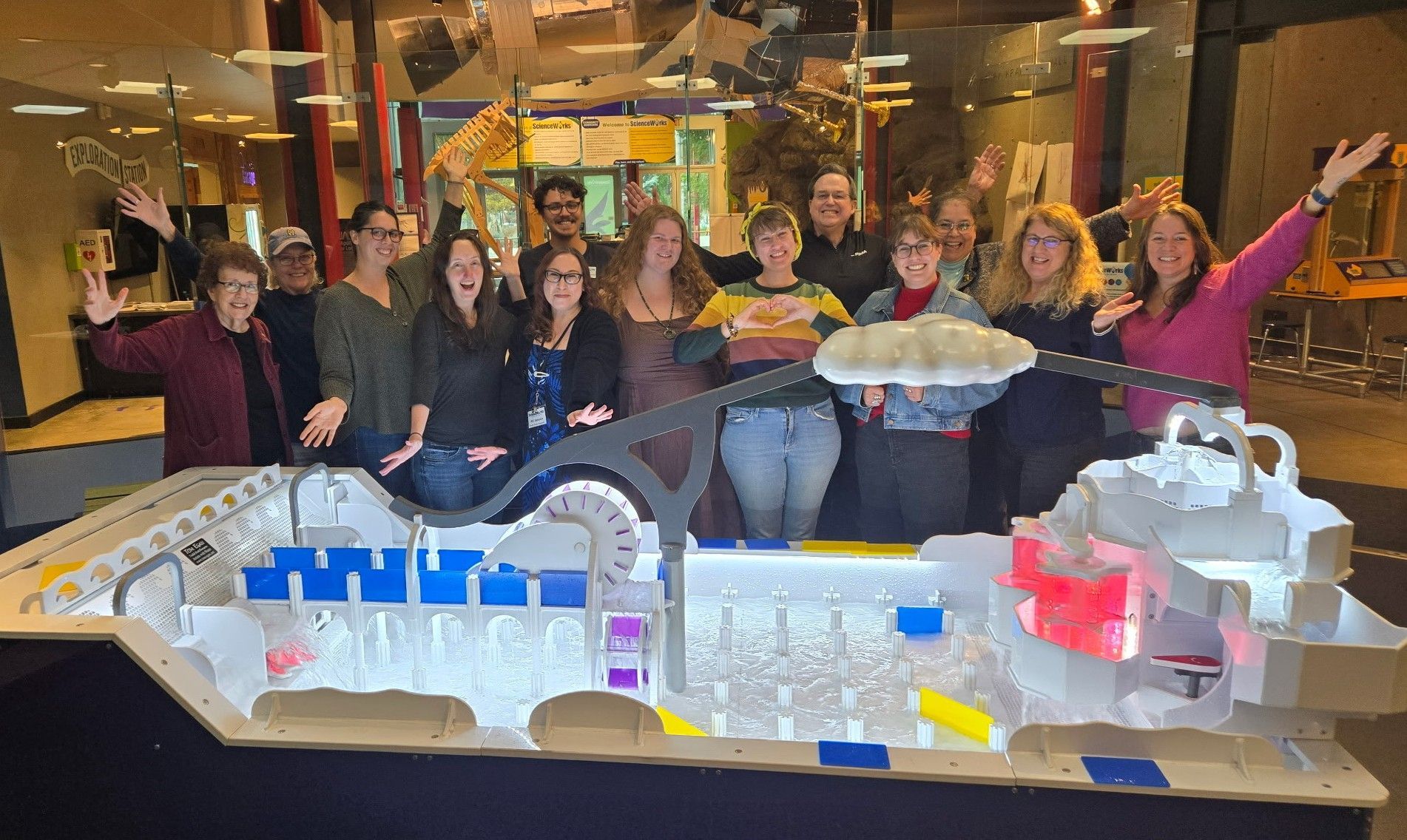 Group of people posing behind the new water table exhibit at ScienceWorks, arms raised. Building interior with colorful decor.