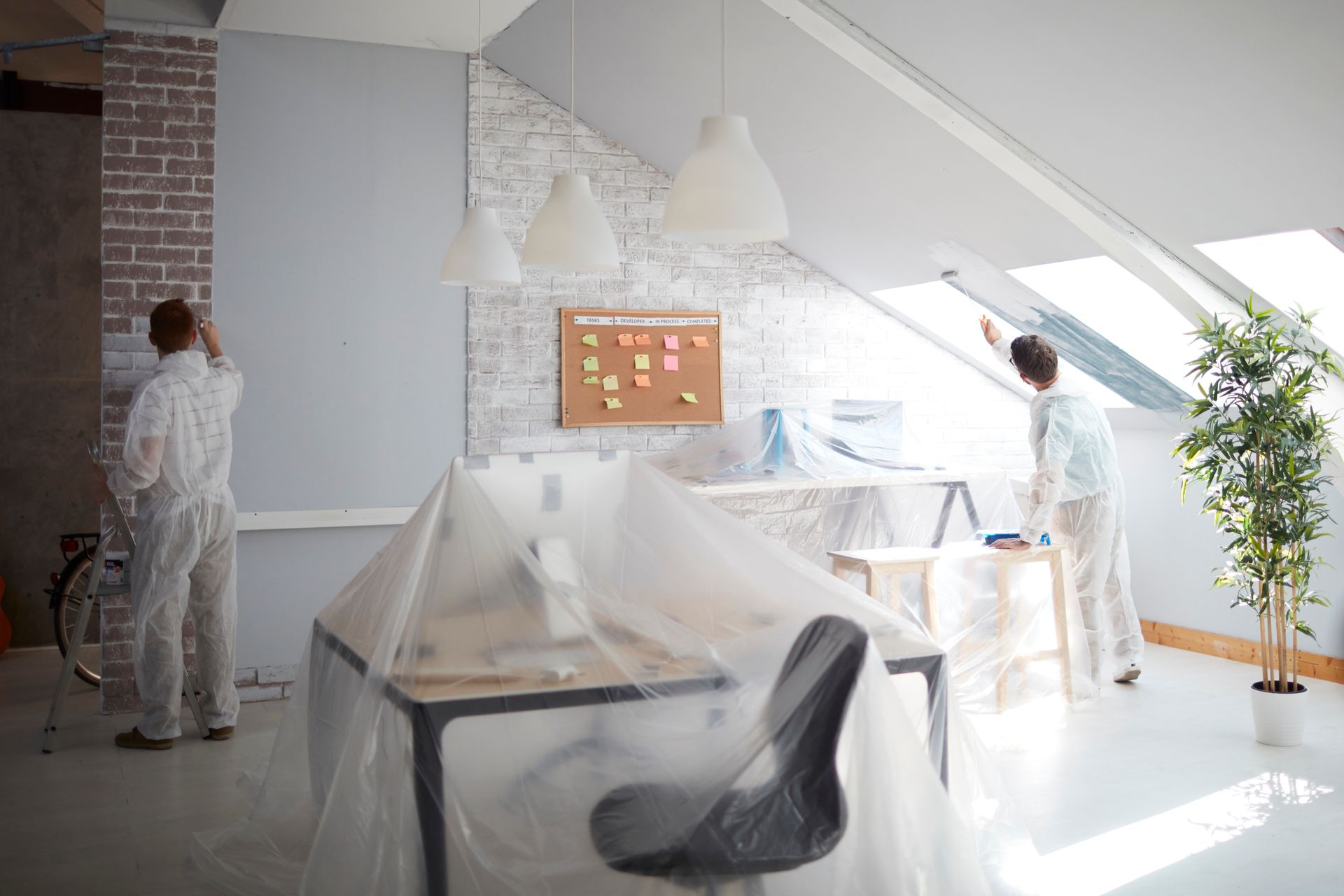 Two workers in protective suits painting a modern office space with plastic-covered furniture.