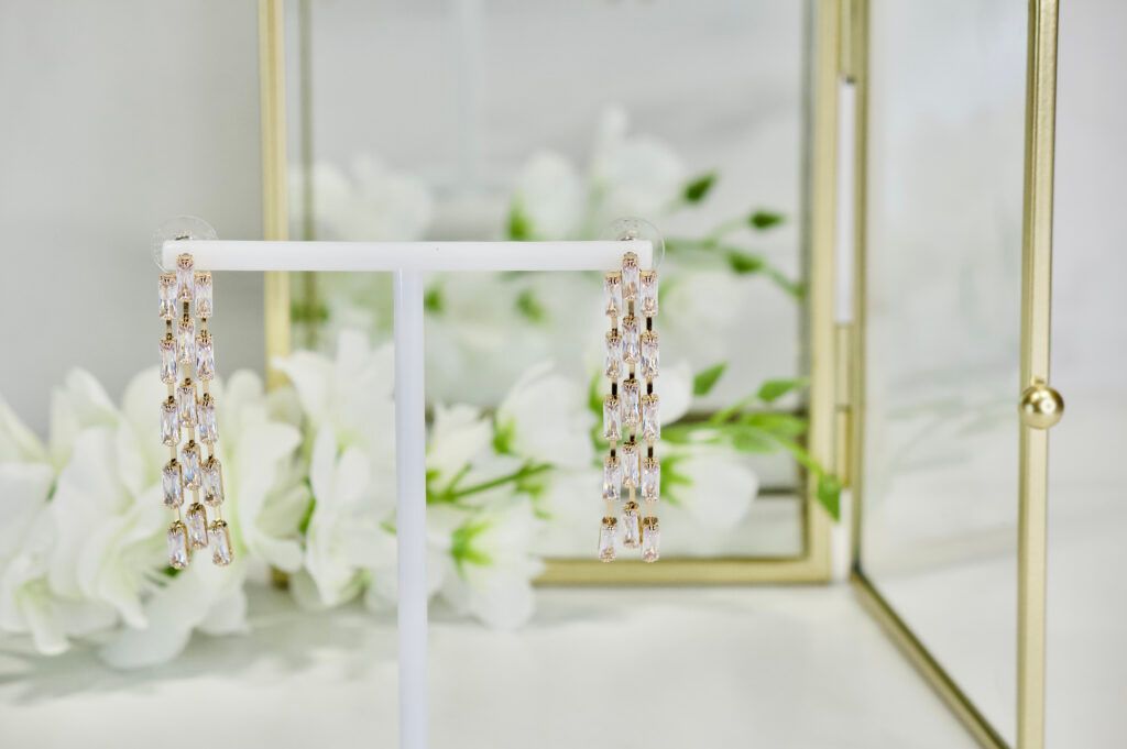 Dangling crystal earrings on a white stand in front of white flowers.