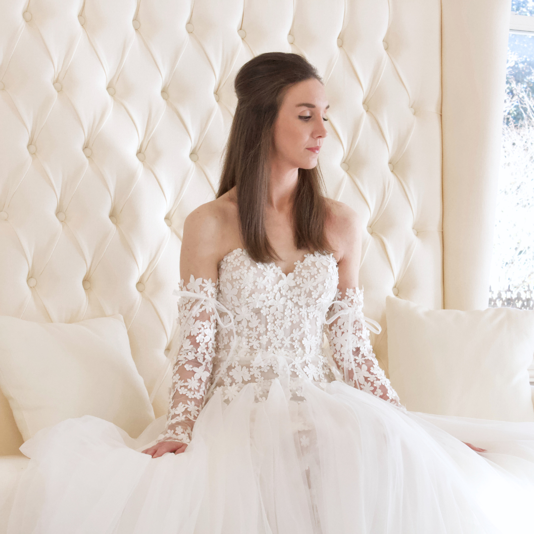 Bride in white lace gown, sitting on a white bed, with a tufted headboard, and a window in the background.