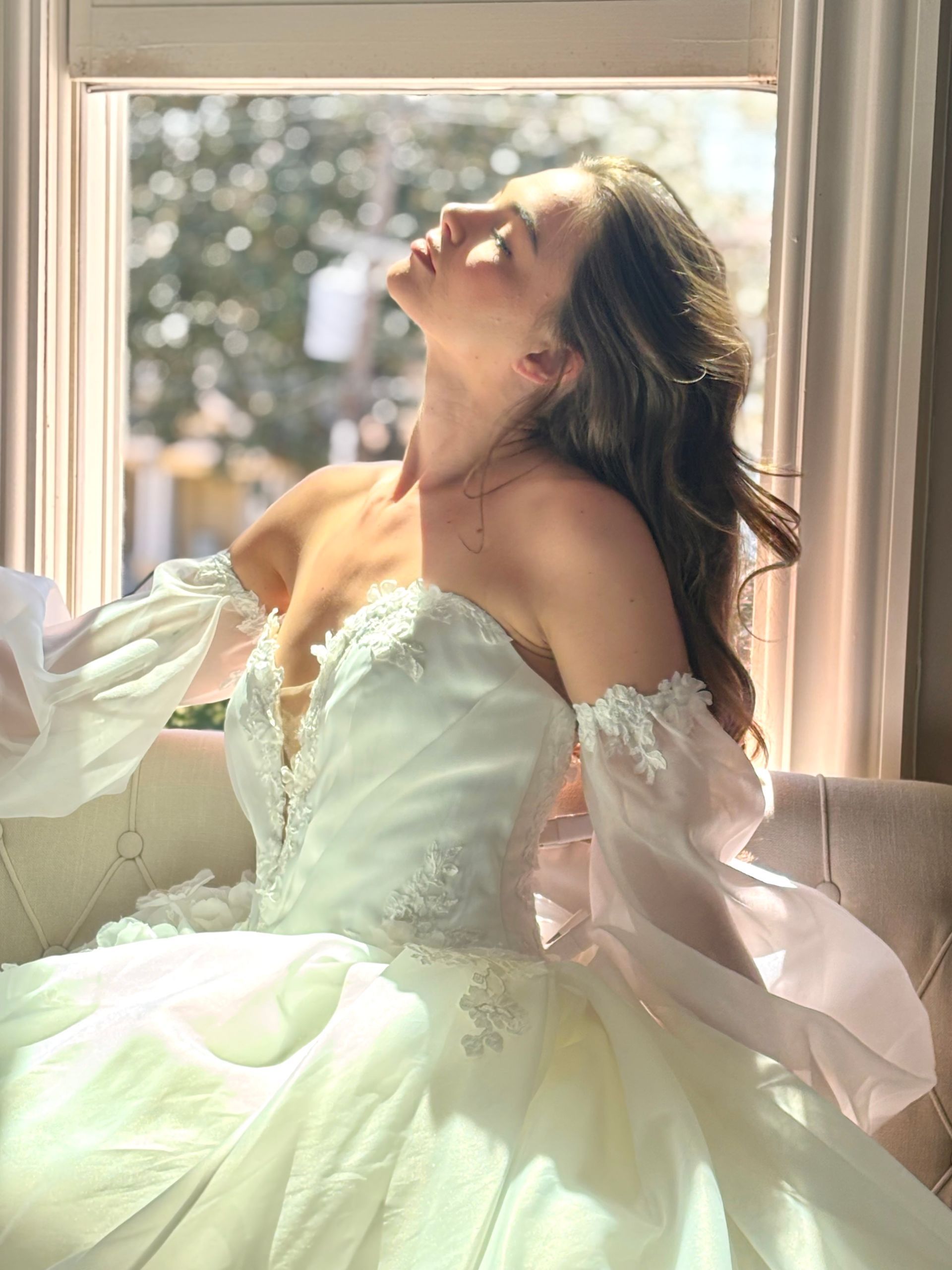 Bride in white lace off-the-shoulder wedding dress, veil, smiling, outdoor setting.