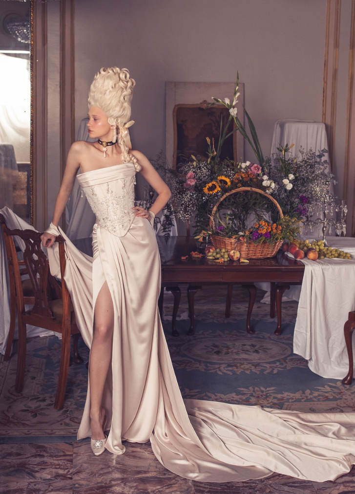 Woman in white period dress, elaborate wig, poses near table with fruit and flowers in a room with ornate decorations.