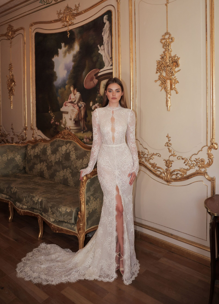 Woman in lace wedding dress with high slit, standing in ornate room with antique sofa and painting.