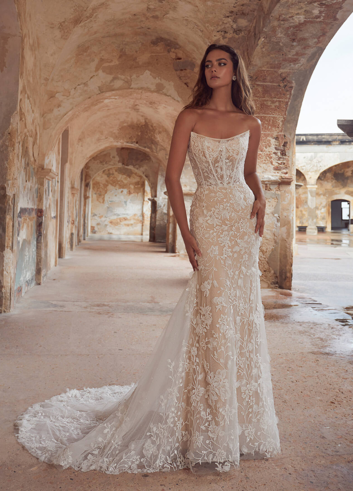 Woman in strapless, embellished wedding dress with train, standing in an arched stone walkway.