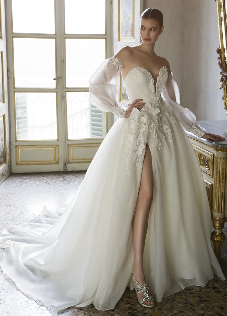 Woman in a white wedding dress with a high slit and long, sheer sleeves. She is standing in a sunny room.