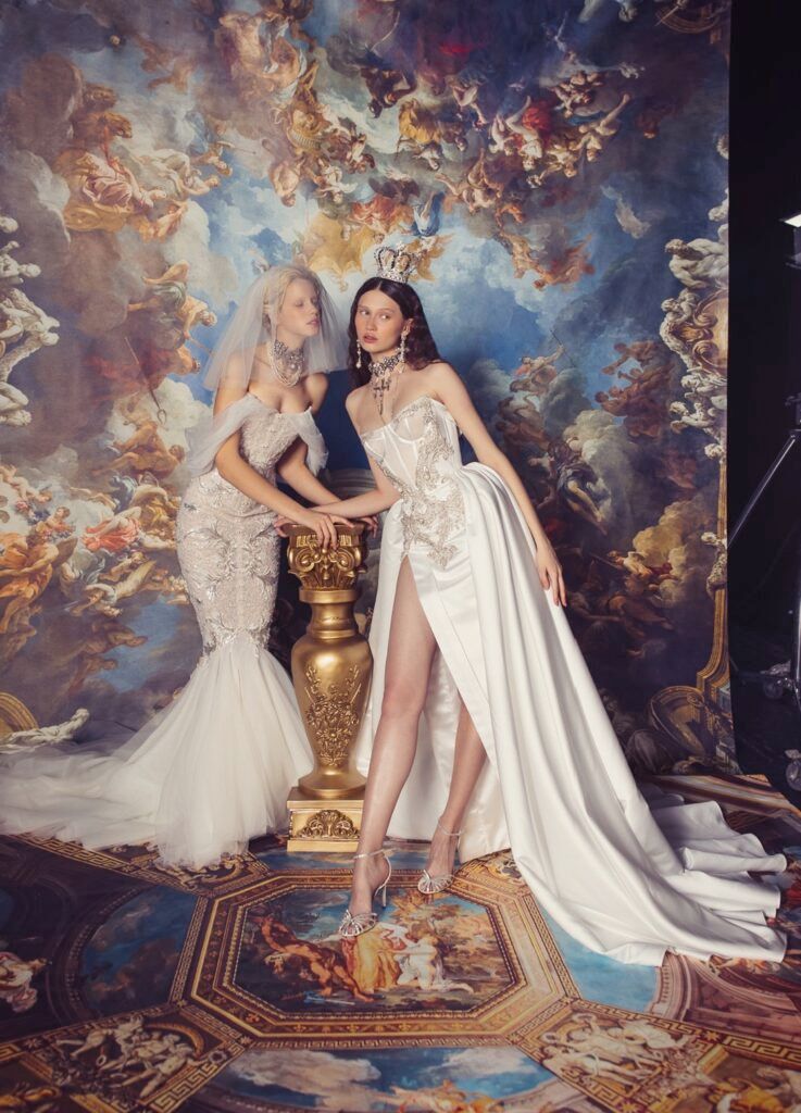 Two women in wedding dresses pose by a golden urn; ornate painted backdrop depicts sky with angels.
