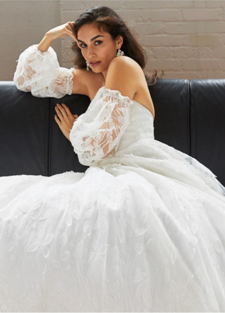Woman in white off-shoulder dress, puffy lace sleeves, sitting on black couch, looking at the camera.