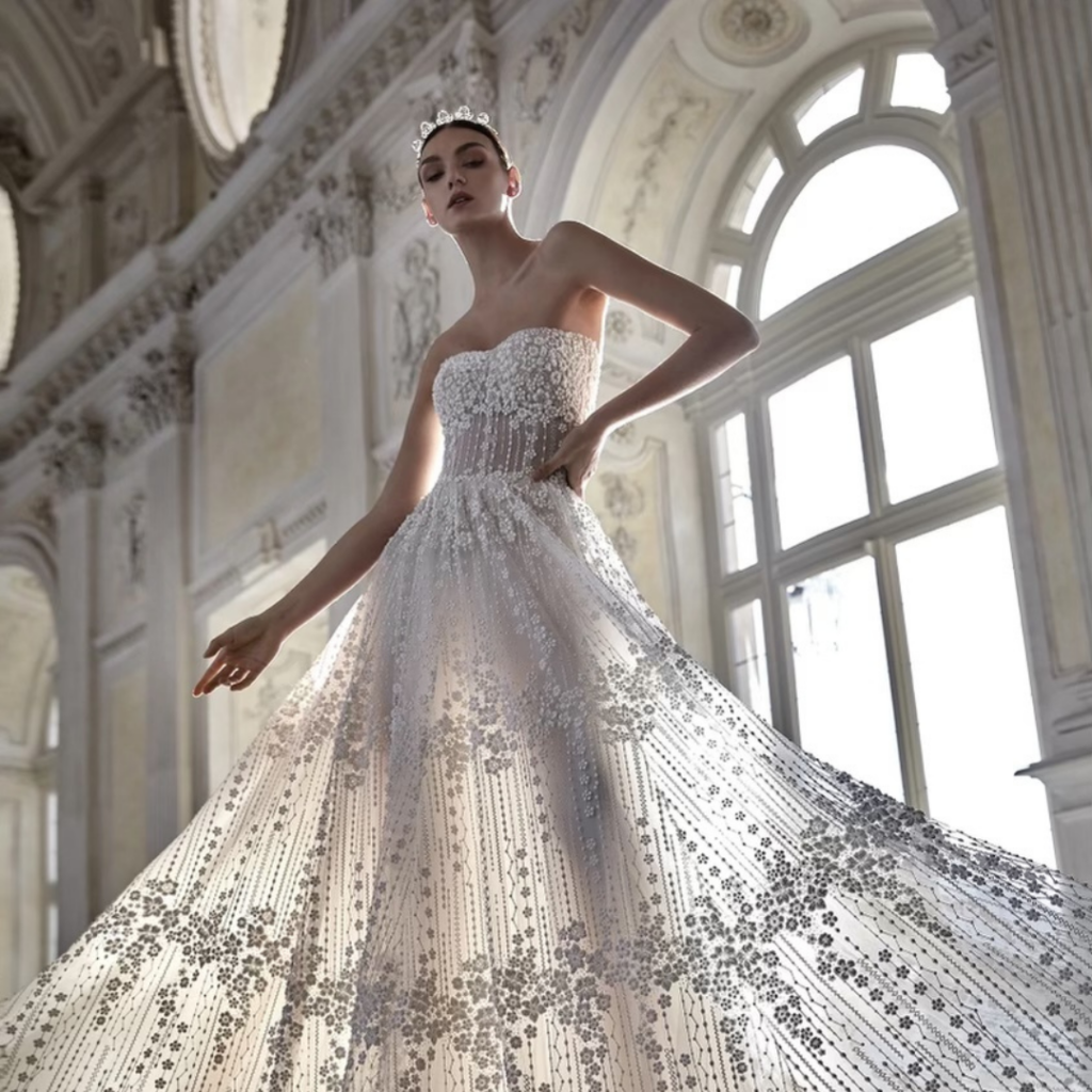 Woman in strapless, embellished gown with tiara, posing in a grand, light-filled hall.