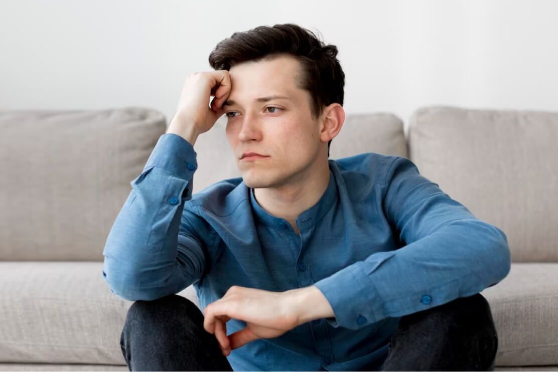 A man in a blue shirt is sitting on a couch with his hands on his head.