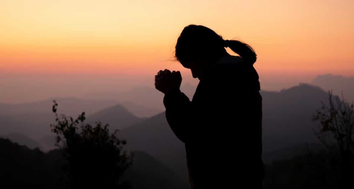 A silhouette of a woman praying with her hands folded in front of a sunset.