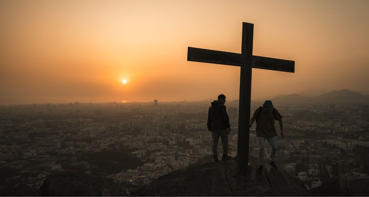Two people are standing under a cross on top of a hill at sunset.