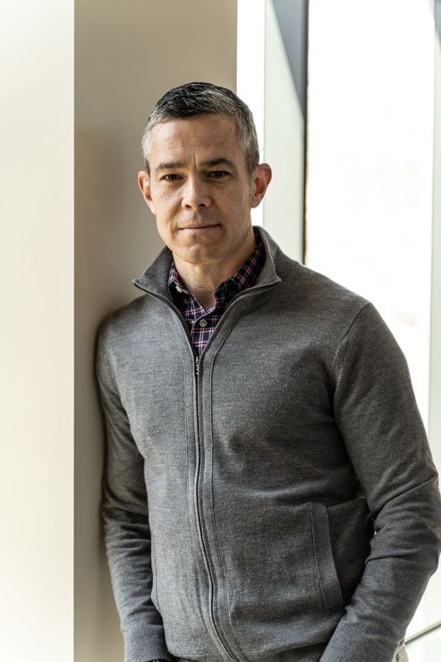 A man in a grey sweater is leaning against a wall.