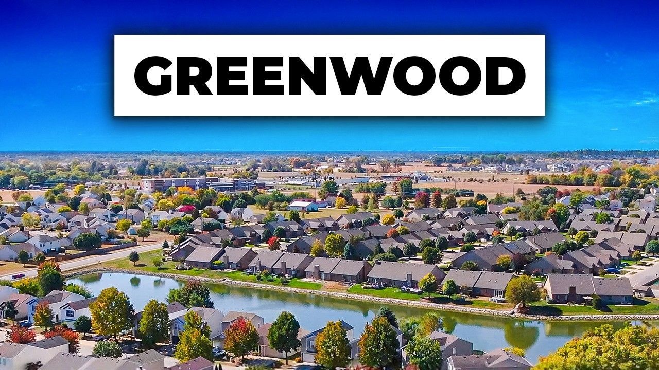 An aerial view of a suburban neighborhood in Greenwood with a central pond surrounded by trees and houses under a blue sky.