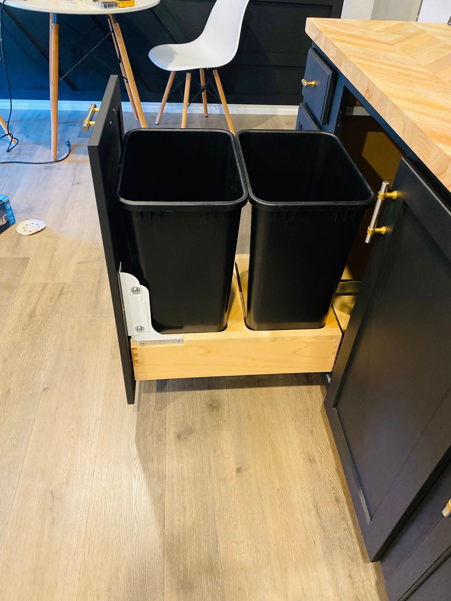 Pull-out kitchen cabinet with two black trash bins. Wooden drawer on wood floor.