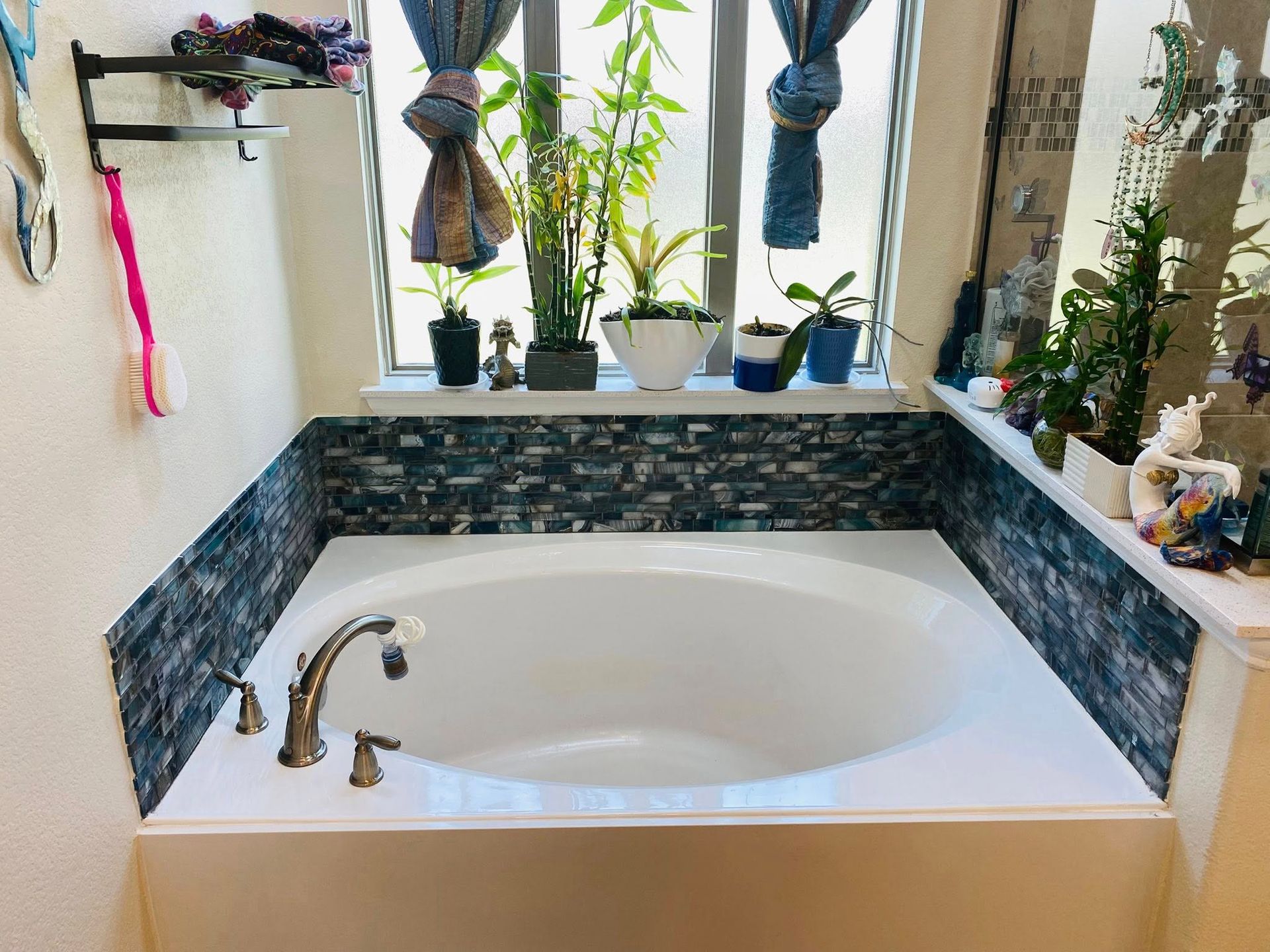 White built-in bathtub with dark tile backsplash, beneath a window with plants and blue curtains.