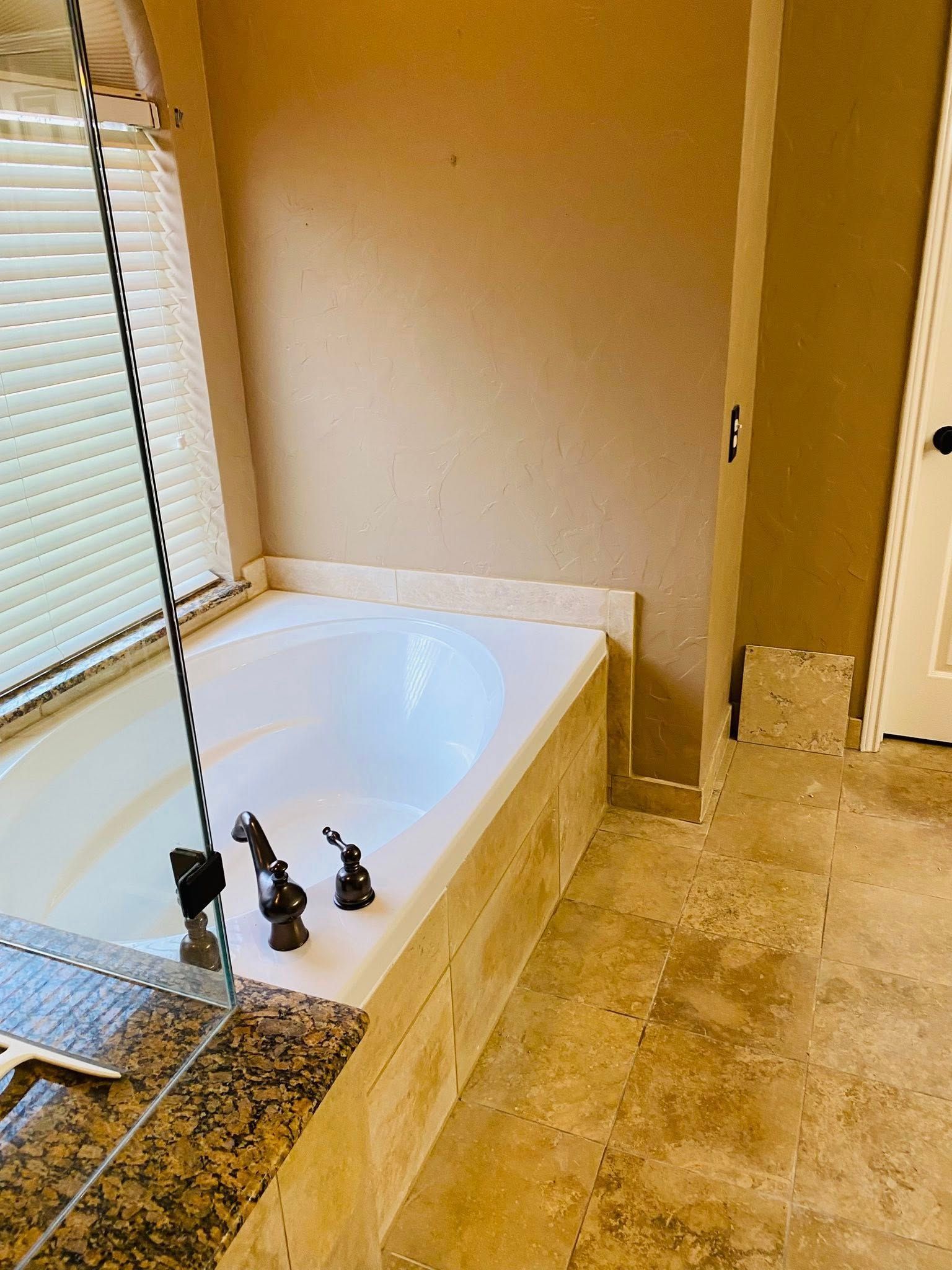 Bathtub in a bathroom with beige tile and tan walls, near a window with blinds.