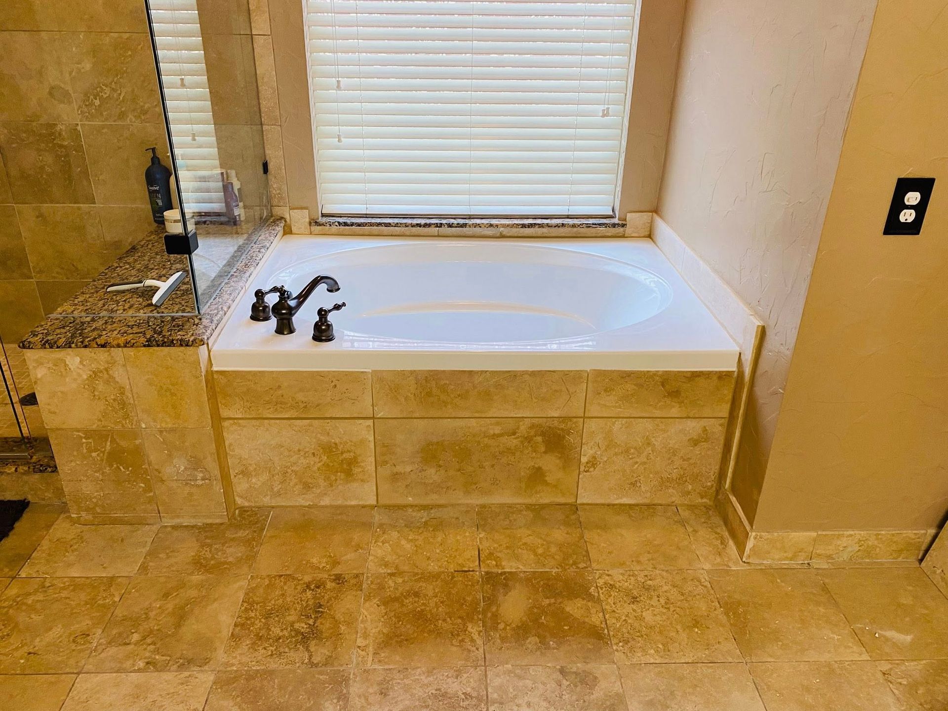 Bathtub in a bathroom with beige tile and a window with blinds. Black faucet.