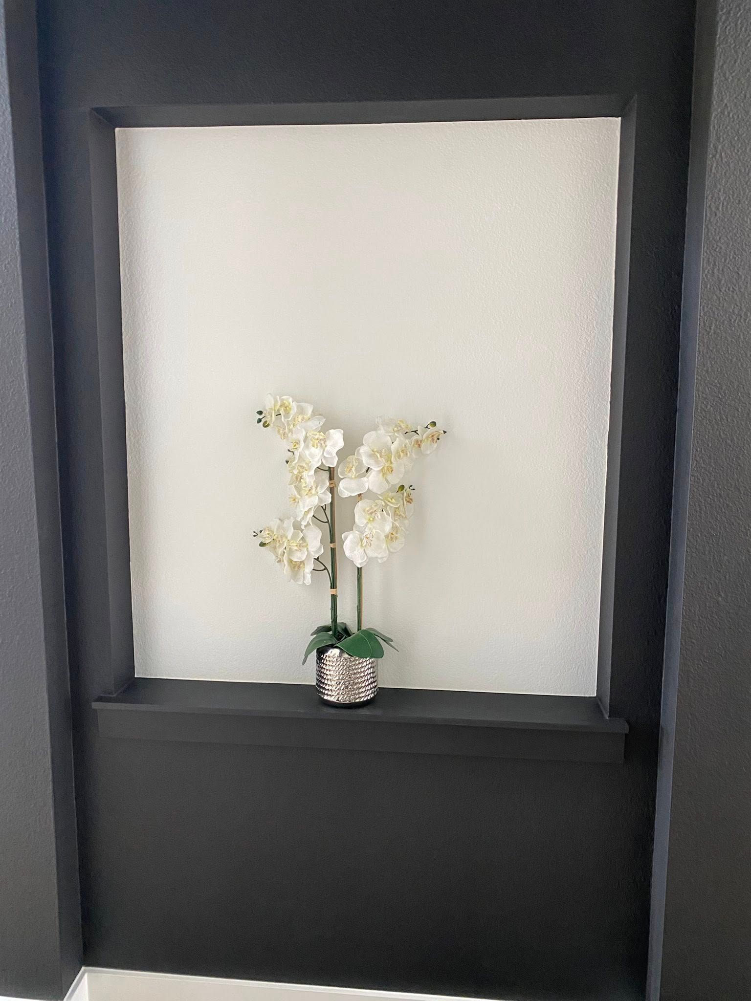 White orchids in silver vase on black shelf in recessed wall niche.