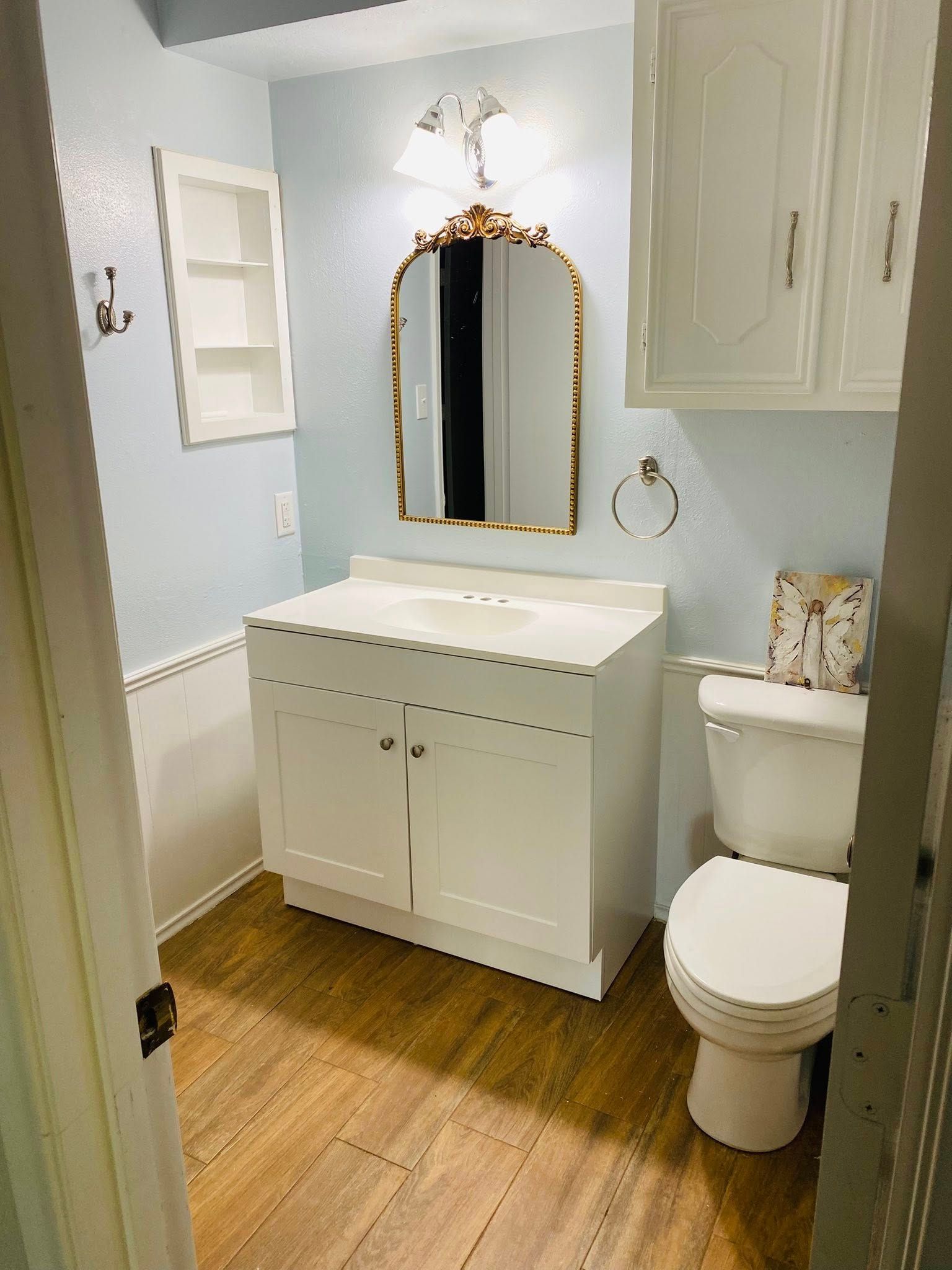 Bathroom with white vanity, toilet, and light blue walls. Gold-framed mirror.