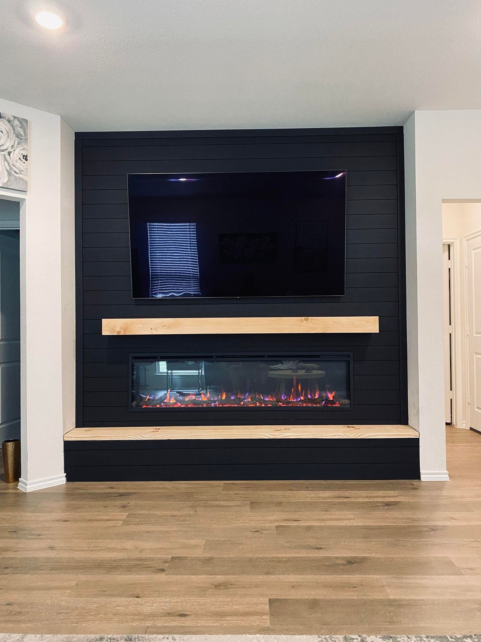 Black accent wall with a mounted TV above an electric fireplace; light wood mantel and hearth.