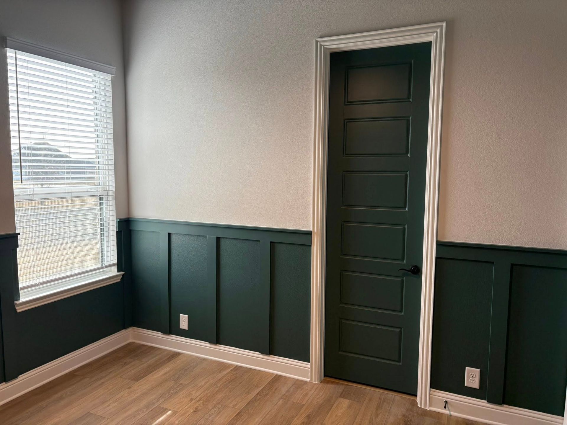 Empty room with green wainscoting and door, white walls, and wood floor. Blinds cover the window.
