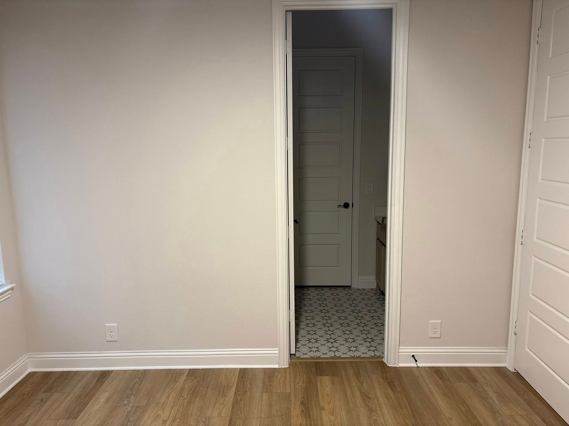 Empty room with light-colored walls, wood-look floor, and doorway leading to a room with patterned tile and a white door.