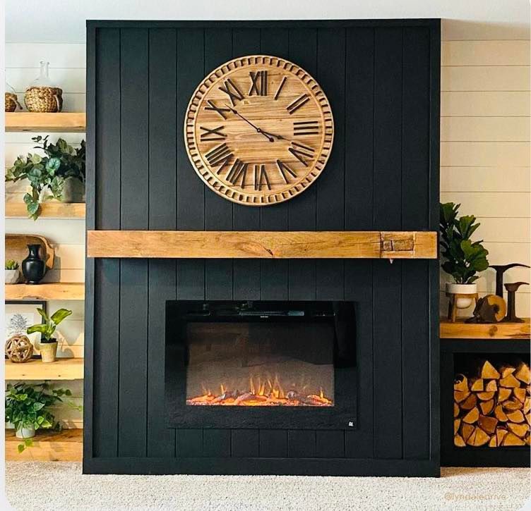 Black fireplace with wooden mantel, clock, and electric fire. Wooden shelves on either side, plants and firewood.