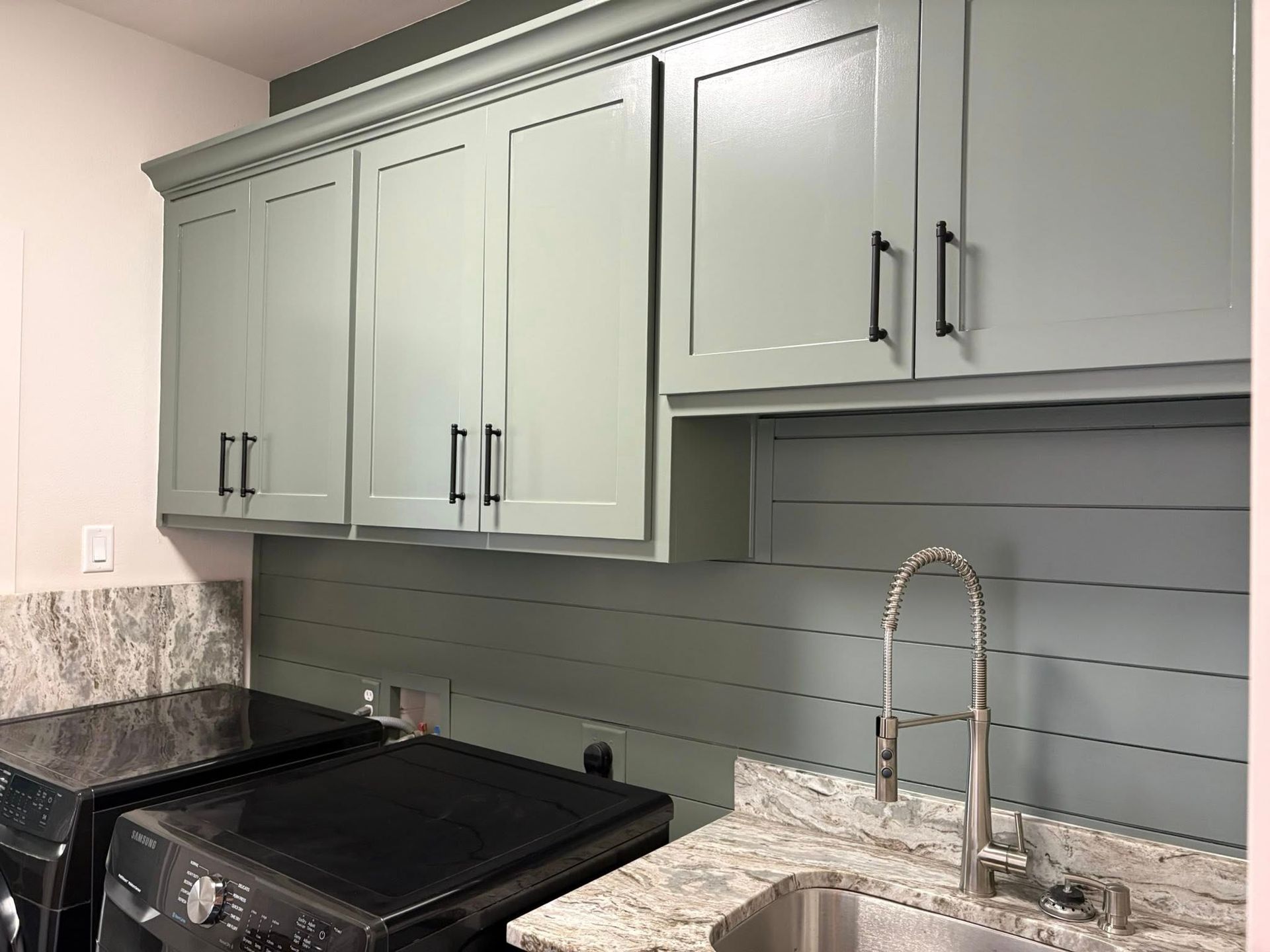 Light green cabinets with black handles in a laundry room, above a shiplap wall and sink.