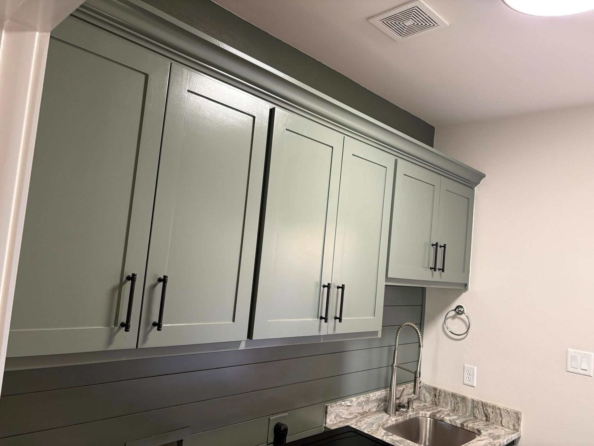 Light green kitchen cabinets with black hardware above a sink and granite countertop.