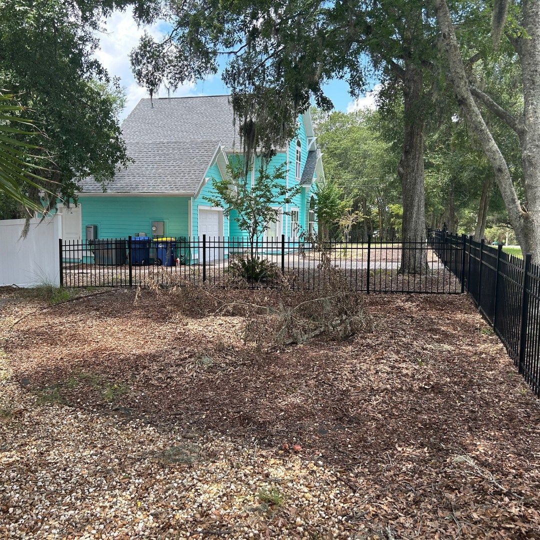 A teal house with a gray roof behind a black fence and brown yard, trees in the background.