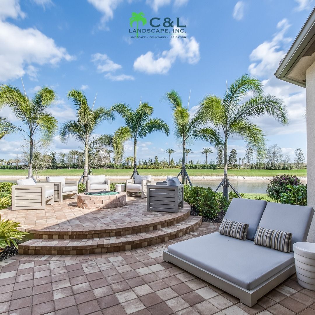 Patio with seating, fire pit, and palm trees, overlooking a lake on a sunny day.