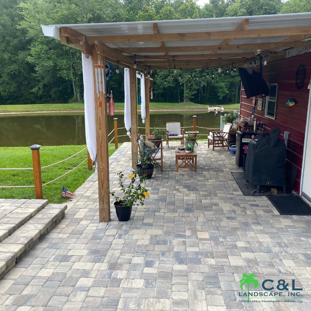 Patio with seating, a grill, and a covered area, next to a pond, in a rural setting.