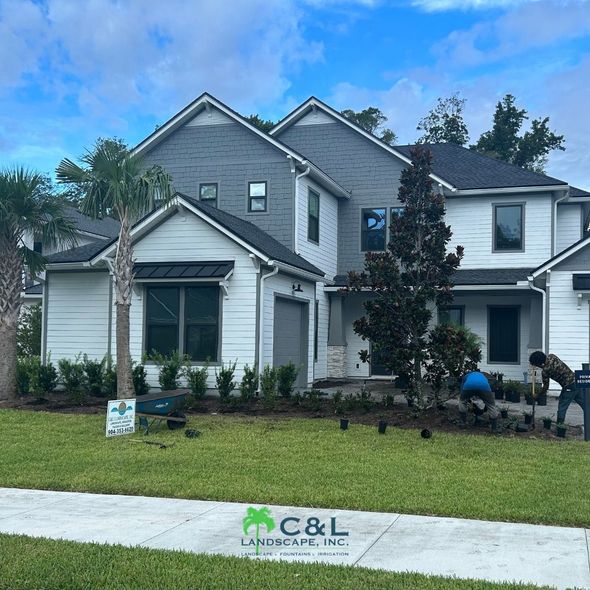 Landscapers planting in front of a white and gray house with green lawn and blue sky.