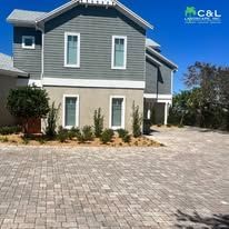Two-story gray house with cobblestone driveway, surrounded by small plants and trees on a sunny day.