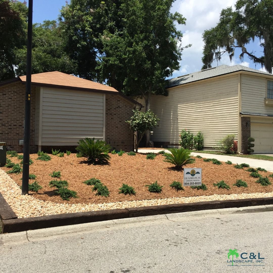 Landscaped yard with wood chips, plants, and a sign. Houses in the background.