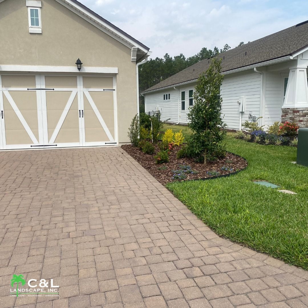 Brick driveway with planted landscaping, garage, and white house on a sunny day.
