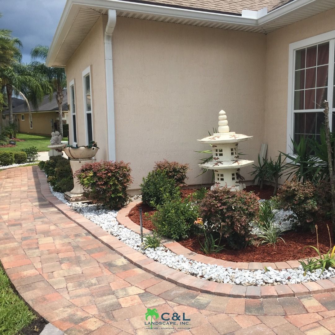 Brick pathway curves past a tan house and garden beds with white rocks, bushes, and a pagoda statue.