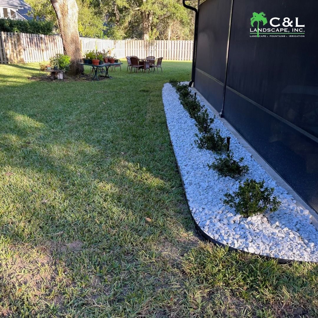 A backyard with a white stone flower bed and green grass. Fence and patio in the background.