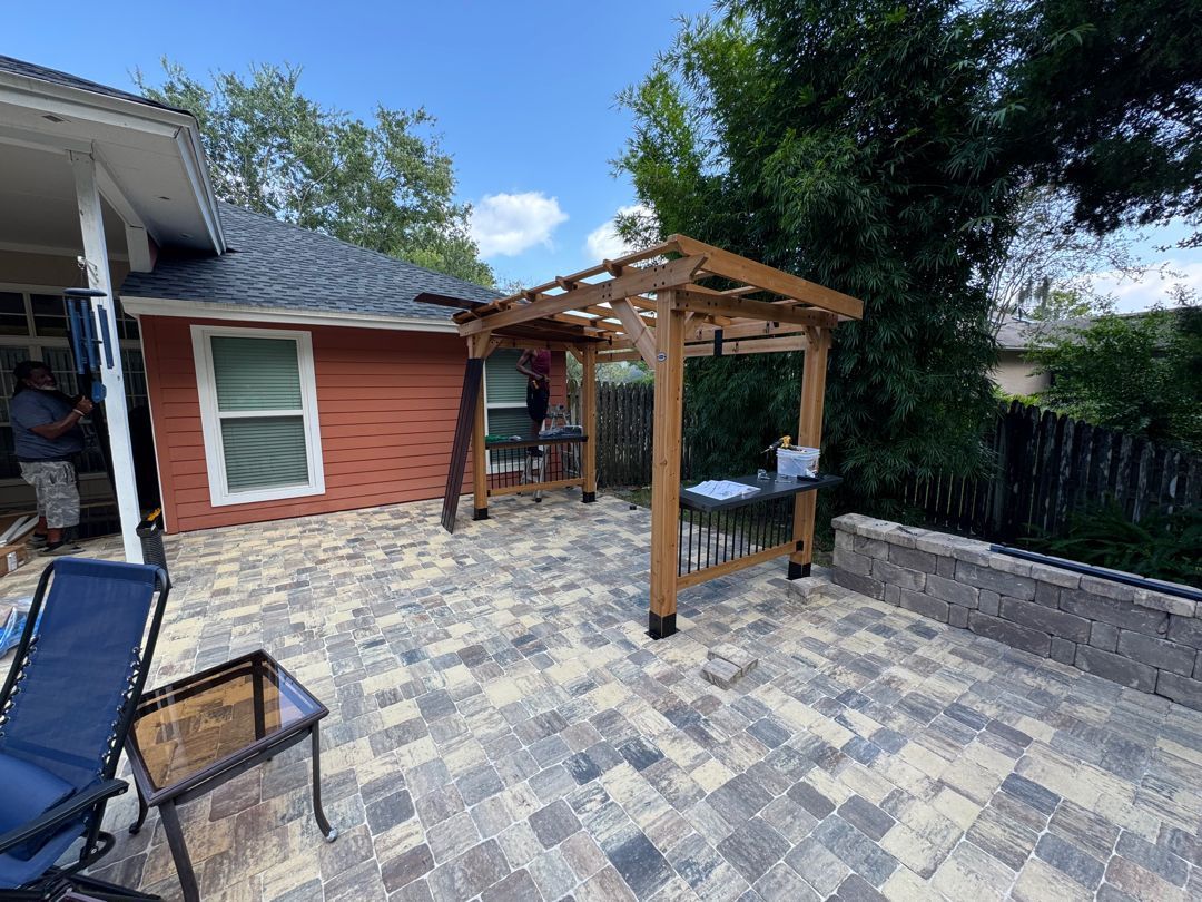 Patio with pergola, brick pavers, blue chair, and red building with a window, under a blue sky.
