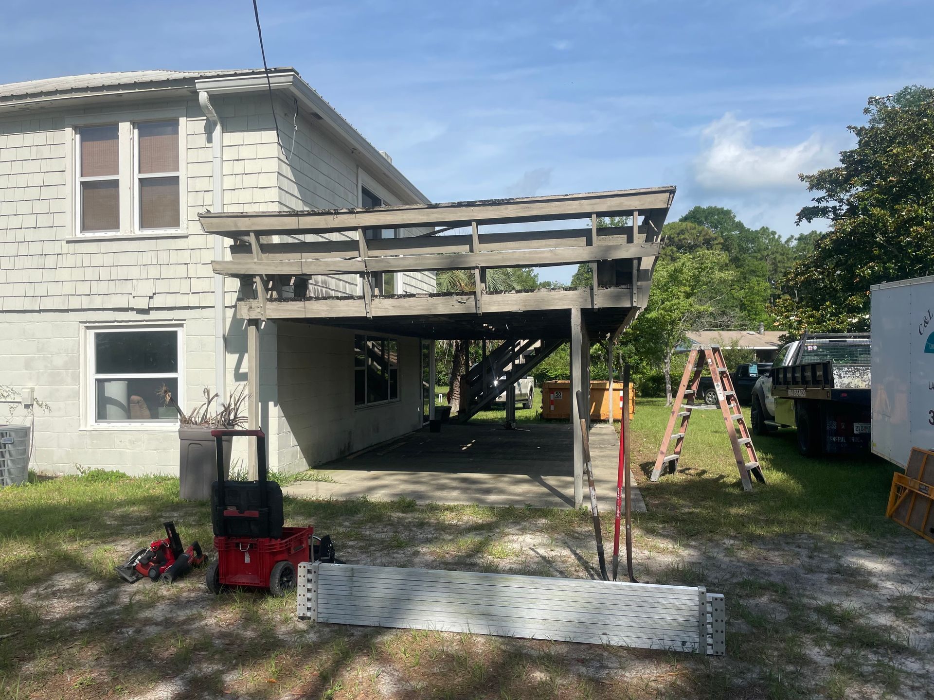Two-story building with a carport under construction; tools, ladder, and materials are visible. Sunny day.