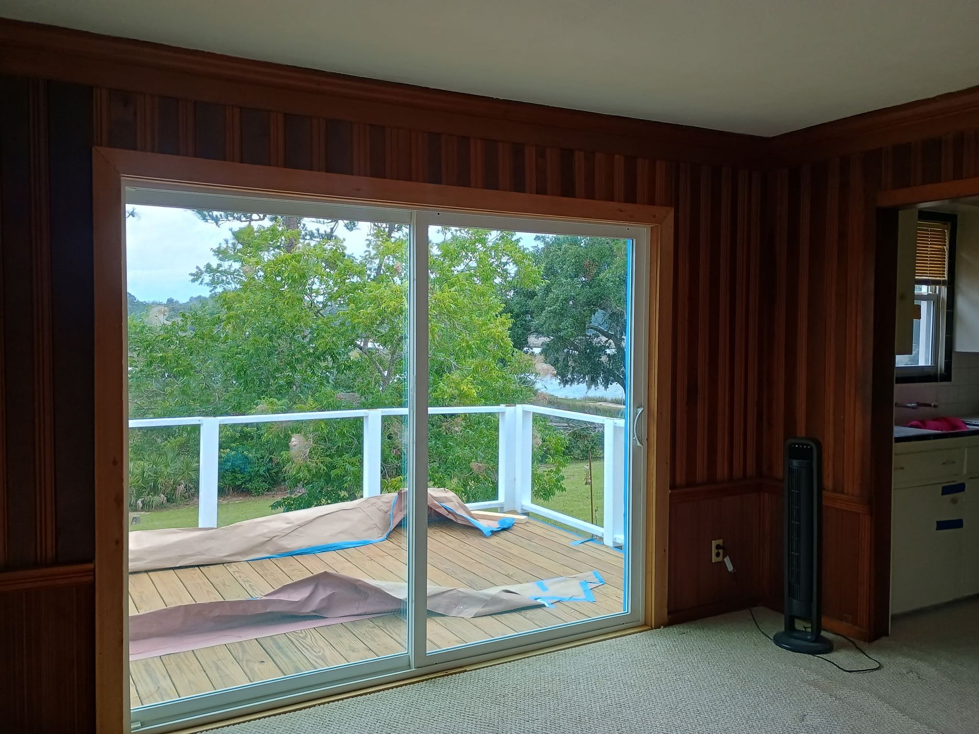 Sliding glass door opens to a deck with trees and a lake view. Wood-paneled walls and a fan.