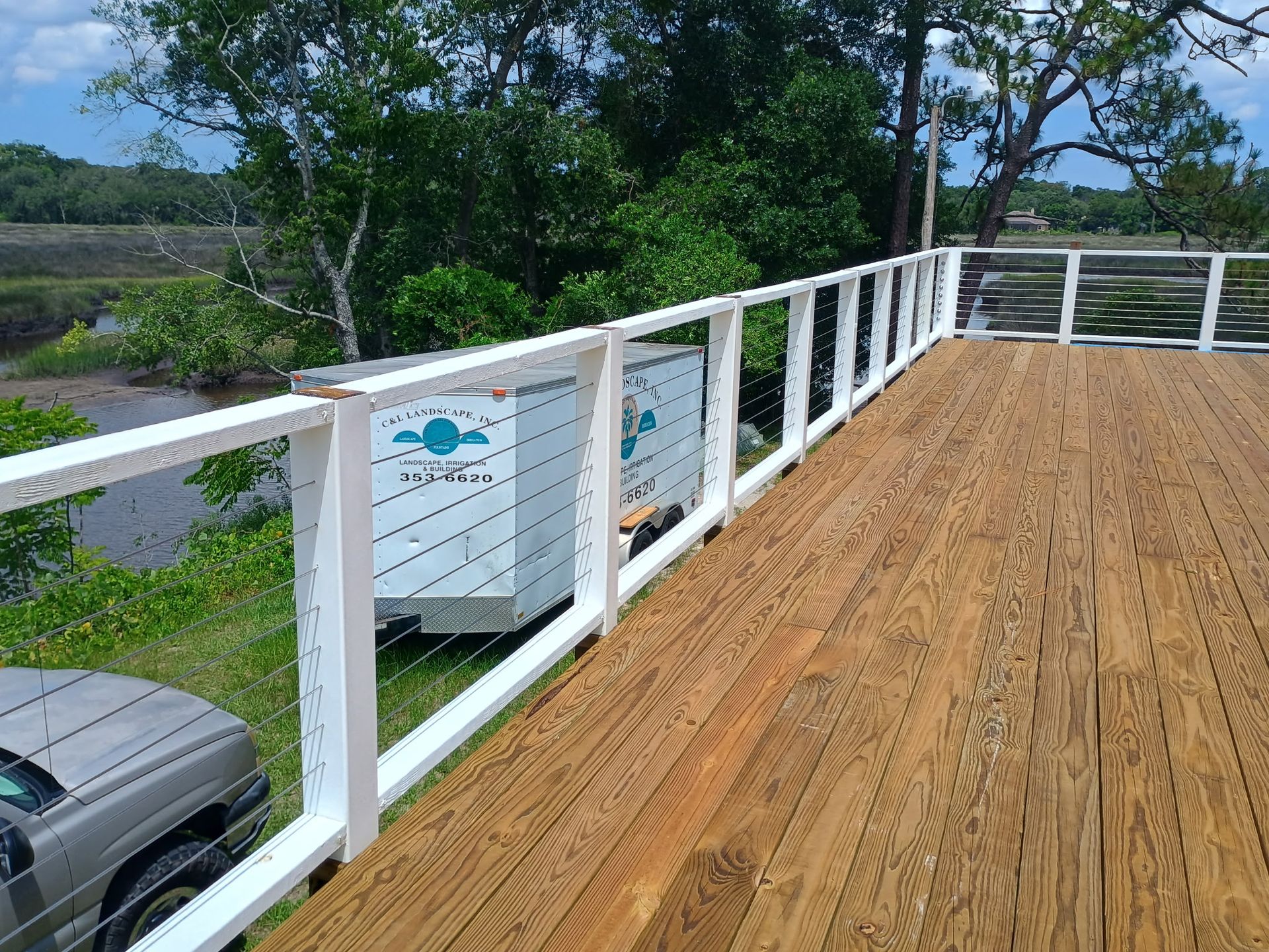 Wooden deck with white railing, overlooking a marsh and trees. A car is parked nearby.