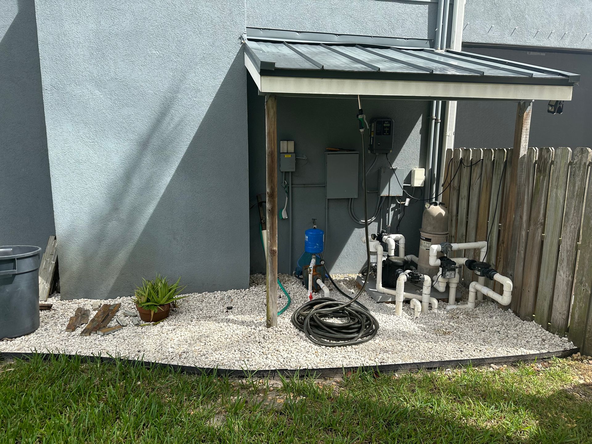 Pool equipment under a small gray awning with gray gravel, a green plant, and a black hose.