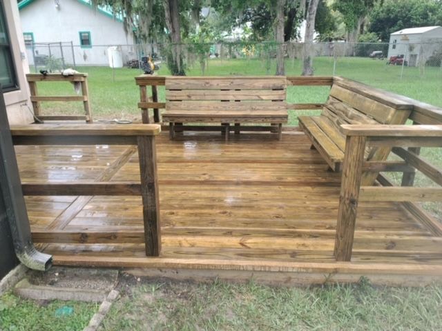 Wooden deck with built-in benches, railings, and a small table. Outdoor setting with grass and trees.